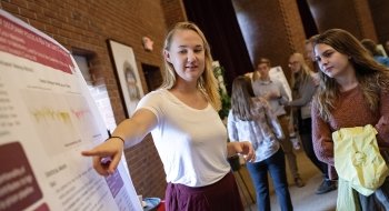 A photo of a student pointing at their research poster and presenting to an attentive listener.