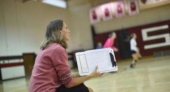 A photo of Shelly Roiger sitting on the sidelines of a basketball court with a clipboard in hand as she shouts out to players.