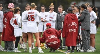A photo of the women's lacrosse team huddled together in a meeting on the field, coach Hannah Corkery is crouching down, the back of her coat reading St Lawrence University.