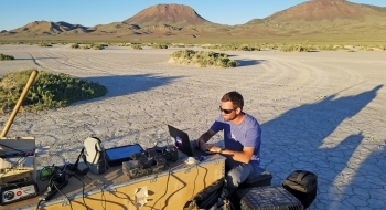 A photo of a student set up and working at a desk in the desert that has multiple devices plugged in.