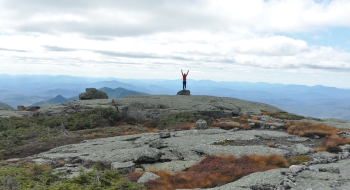 A photo of a person on top of a mountain with their arms up and outstretched over a vast landscape.
