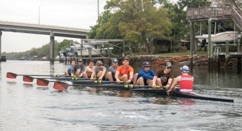 The men's crew team row together on a boat on a river in Tampa Bay on a warm, overcast day.
