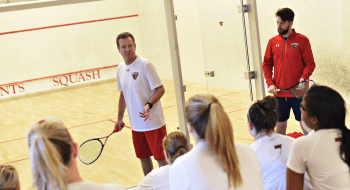 A photo of women's squash coach Scott Denne demonstrating something to the team who are sitting and watching.