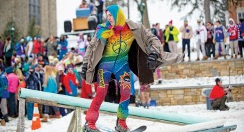 A photo of students at rail jam in brightly colored clothes. In the foreground is a person in rainbow clothes skiing on the rail as everyone behind him watches.