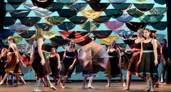 A photo of St Lawrence dancers performing on stage. They are all wearing black dresses and are dancing in front of a wall full of umbrellas.