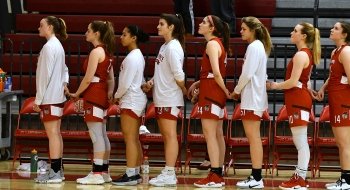 A photo of members of the women's basketball teamed lined up, with each athlete holding the pinky fingers of the people ahead and behind them.