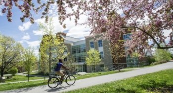 A student riding a bike on a sunny spring day on campus with a stone building in the background