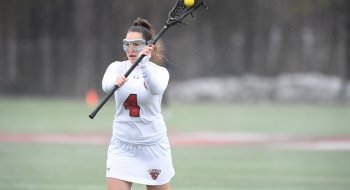 Nicole Stanton, wearing a white Saint Lawrence University lacrosse uniform, cradles the ball and prepares to pass during a home game.