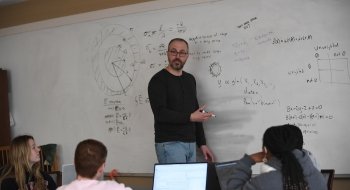 Professor Ivan Ramler stands at the front of a class explaining an equation on a whiteboard.