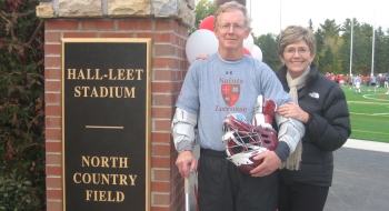Don and Sue Martin stand in front of Hall-Leet Stadium at North Country Field on the Saint Lawrence University Campus. Don is wearing a grey Saints Lacrosse shirt and holding a lacrosse helmet and stick as a lacrosse game takes place in the background.