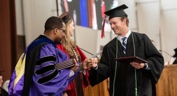 A Saint Lawrence graduate, wearing a black cap and gown, is congratulated by President Kathryn Morris and Hagi Bradley on the Commencement stage.