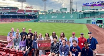 A group of students holding a Saint Lawrence University flag stand in the stands in front of the field at Fenway Park.