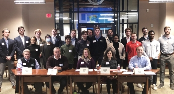 A large group of Saint Lawrence University students stand behind a table of alumni judges.