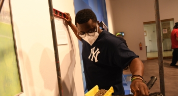 Michael Yirenkyi '22, wearing a New York Yankees t shirt, reaches for a hammer while installing his art piece on a gallery wall.