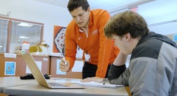 Eduardo Puerta, class of 2022, wearing a red quarter-zip sweatshirt, holds a marker while explaining a math problem to a fellow student.