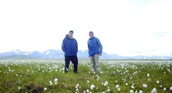 Jon Rosales and a fellow research, wearing royal blue rain jackets, stand in a field of green grass and white flowers. Large snowcapped mountains are in the distance.