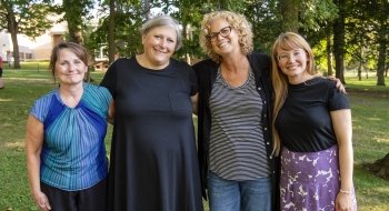 Four faculty and staff members gather for a group photo in a bright wooded area on campus.