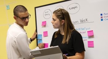 Blake Bittorf stands at a whiteboard with writing and sticky notes on it. He and his colleague look at her computer while reviewing a plan for social media.