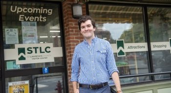 Charles Shaw, wearing a checkered button-down shirt, stands in front of the front door of the Saint Lawrence County Chamber of Commerce.
