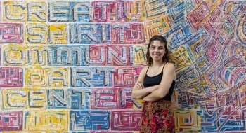 Mikayla Quinn, wearing a black tank top and patterned skirt, stands in front of a colorful mural at a community art center.