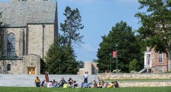 A professor teaches a class outdoors on a beautiful late summer day on the quad, framed by a stone chapel and brick academic building.