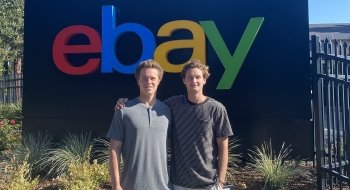 Two college-aged men wearing casual clothing stand in front of the eBay sign.