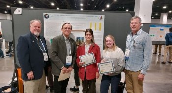 Two female college students hold award certificates and stand in front of their research poster at a geology conference in a large hall. Their professor and two judges stand next to them.