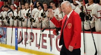 Joe Marsh, wearing a scarlet blazer, waves to the crowd in Appleton Arena as he steps out onto the ice. The Saint Lawrence men's ice hockey team is standing near the bench in the background.