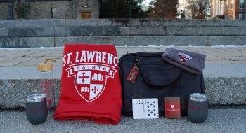 A photo on the steps from the top of the quad with two gray St. Lawrence tumblers, a large red blanket with the St. Lawrence Saints logo with the crest in white, a leather bag, a St. Lawrence deck of cards with a royal flush in spades revealed and a gray tuc with the St. Lawrence Saints logo.