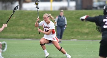 Greta Massey, wearing a white Saints Lacrosse jersey, cradles a lacrosse ball as she moves down the field against the competition.