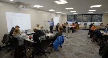 Michael Shuckers stands at the front of a classroom and points to a lesson on a marker board.