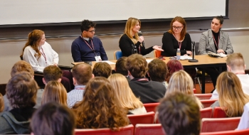 Five panelists sit at a table, facing an audience of students in theatre-style seating. The panelist in the center is speaking with a microphone in one hand, with the other hand is held out.