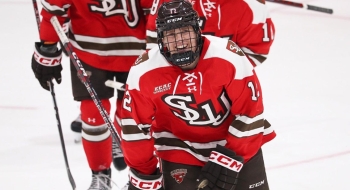 Hockey player Greg Lapointe in a red St. Lawrence University jersey and full hockey pads, smiles while skating on the ice at Appleton Arena.