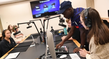 A group of students learn how to use microphones and audio equipment in a studio designed for podcasting. One student stands up and manages controls while others look on.