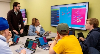 A group of four students sit at a conference table. Their laptops are open in front of them as they discuss the material on a large screen mounted on the wall next to them. Their professor stands at the table while engaging with his students.