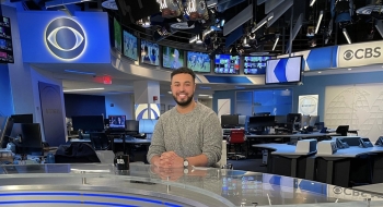 Michael Paulino, wearing a gray sweater, sits at the CBS News Desk in the main newsroom. He's surrounded by blue and white screens. There are two digital CBS logos in the background.