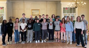 A group of Saint Lawrence students gather together with President Morris in celebration of academic excellence. There is a large gold chandelier hanging from the ceiling of the white room.