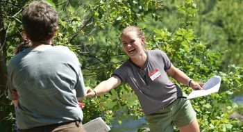 A person wearing a gray shirt and a red name tag, stands outside on a sunny day, holding a white piece of paper hands a small research sample to a community member participating in the Nature Up North Program.