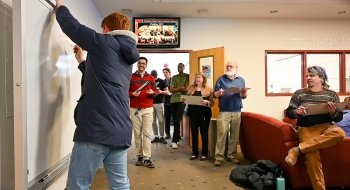 One person is standing at a whiteboard, drawing a circle. A group of seven people sit or stand behind the person drawing, watching the process.