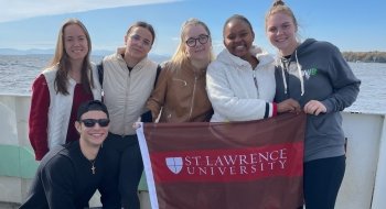 A group of Saint Lawrence University students hold a scarlet and brown flag on a boat.
