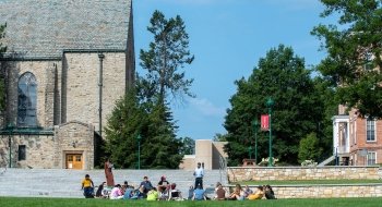 A bright beautiful late summer day on the quad. A professor is teaching a class at the base of stone steps leading up to a tree-lined walkway. In the background is a stone chapel and a brick academic building.