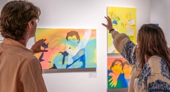 In an art gallery, people hold their hands out in front of their faces while they discuss three pieces of artwork painted in yellow, coral, and blue.