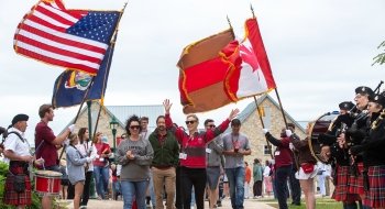 A group of smiling Saint Lawrence alumni participate in a parade for Reunion while bag pipe players play music and members of the community hold American, Canadian, New York, and Saint Lawrence flags billowing in the wind.