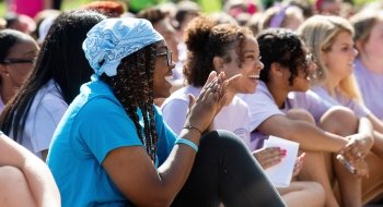 A large group of students in plain blue and lilac t-shirts sit in a group, applauding for a performance out of frame.