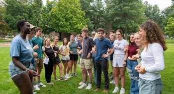 A group of first-year students, lead by their Orientation Leader, link arms while playing ice-breaker games on a grassy clearing.