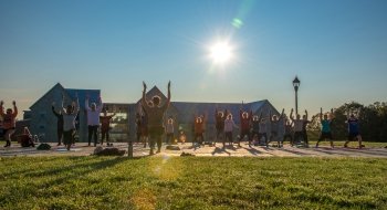 group of students doing yogo on the quad lawn platform, session led by one person. In the background we can see the Kirk Douglas Hall and a beautiful sunrise shining through, onto the students, the building and effecting the image slightly.