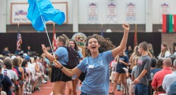 A student, wearing a robin blue t shirt, jumps in the air in excitement while waving a bright blue flag in the air. A sea of Saint Lawrence student in their families sit in chairs inside Newell Fieldhouse in the background.