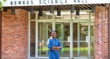 Francesca Mnenula wearing a blue jean dress and standing in front of a mid-century modern brick science building.