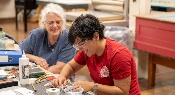 Alejandra paints a small book of paper that she made. The desk in front of her is cluttered with cutting and painting supplies.
