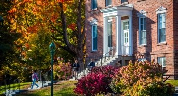 Students walks down the stairs of an early-Italianate style academic building made of red brick, surrounded by fall foliage.